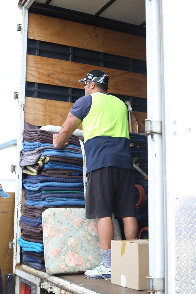 A mover standing inside a truck beside stacked moving blankets and a covered mattress.