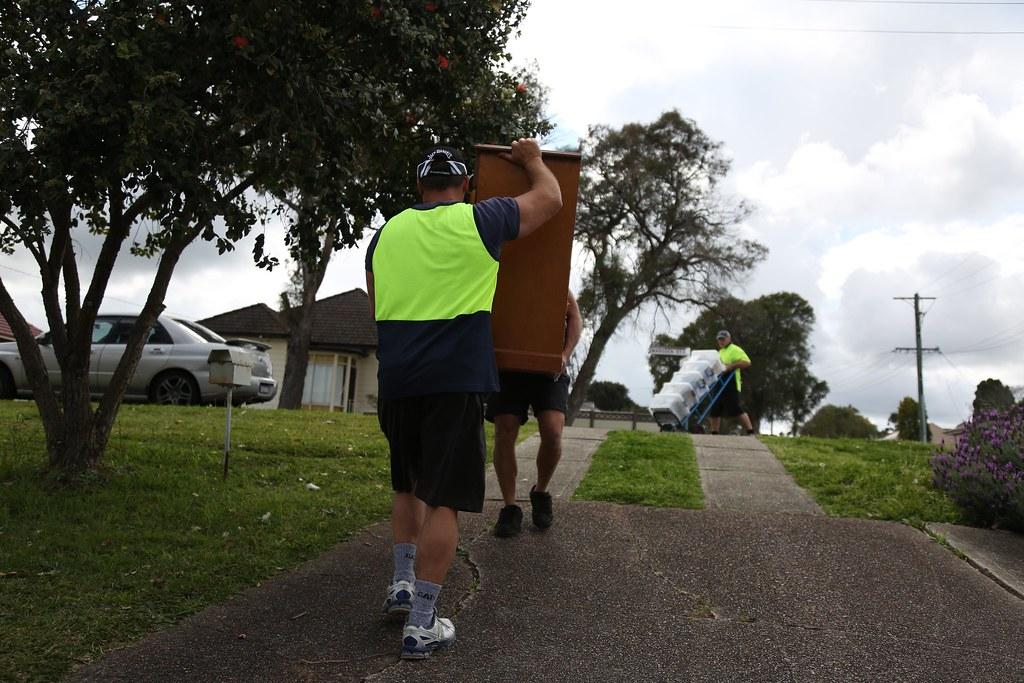 Movers carrying a wooden cabinet along a residential path, with another mover transporting boxes behind them.